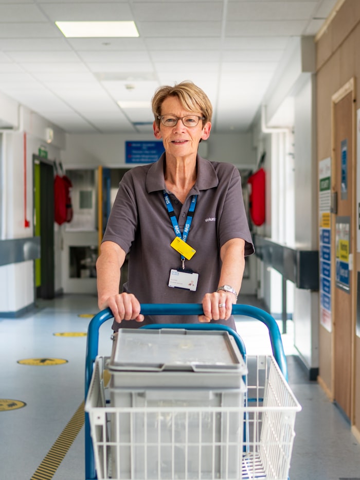 A smiling hospital care worker pushing a supply cart down a ward corridor