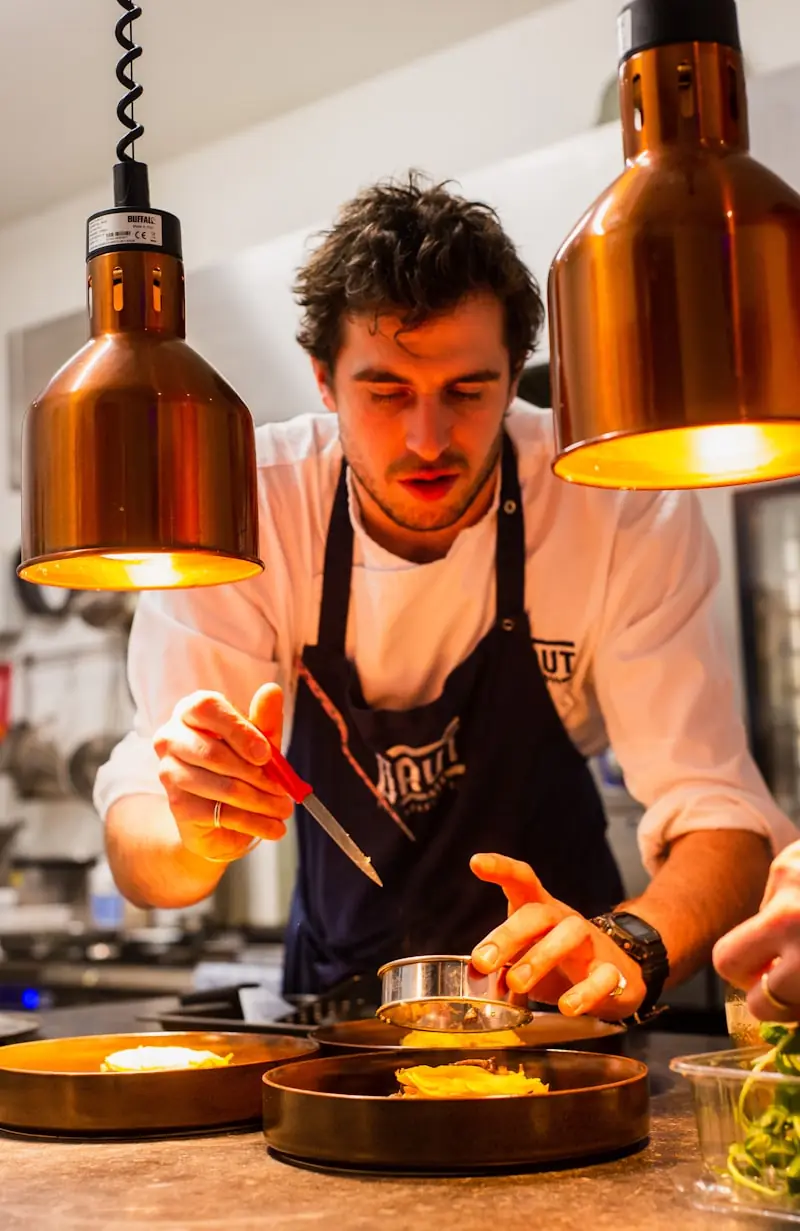 A cook plating a dish under warming lamps in a restaurant kitchen